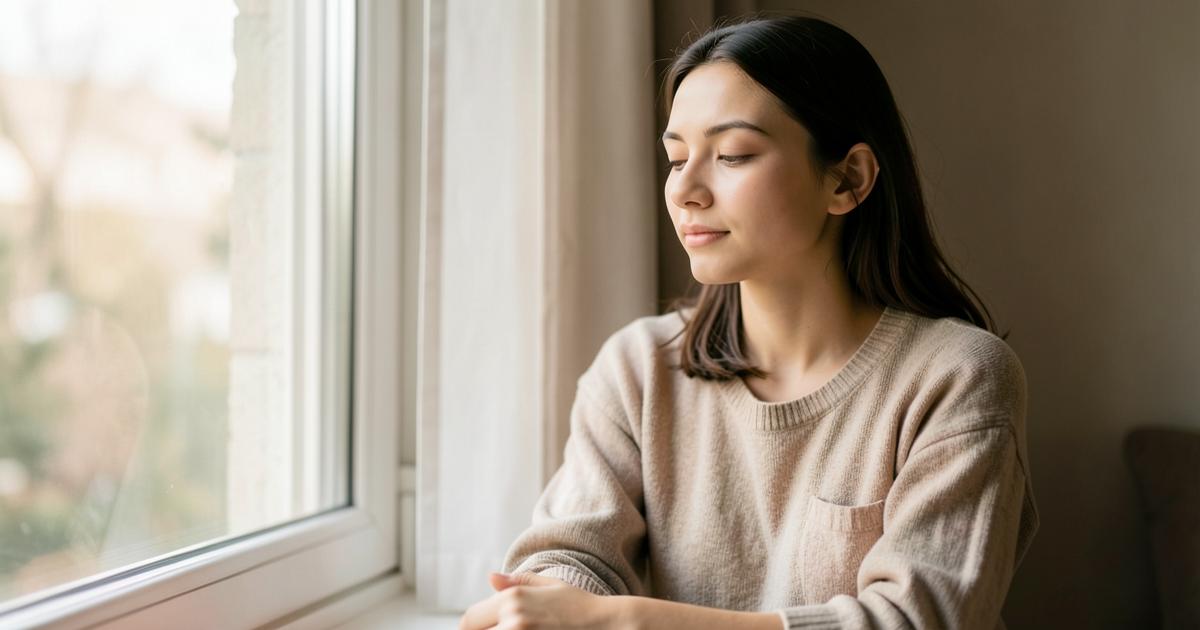Mujer joven sentada junto a una ventana con luz suave, en actitud reflexiva